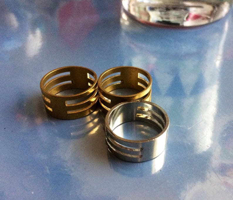 May include: Three metal rings with a slotted design. Two rings are gold-toned, and one is silver-toned. The rings are cylindrical with three horizontal slots. The rings are displayed on a reflective, light blue surface.