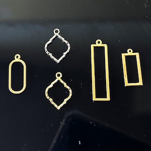 Op de afbeelding: Een assortiment metalen sieradenbedels in verschillende vormen. De bedels omvatten een ovaal, twee vierpassen en twee rechthoeken. De bedels zijn goud- en zilverkleurig, met een kleine lus aan de bovenkant.