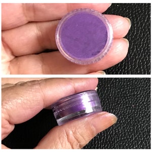 May include: A small clear plastic jar containing a purple iridescent pigment. The jar is being held in a hand.