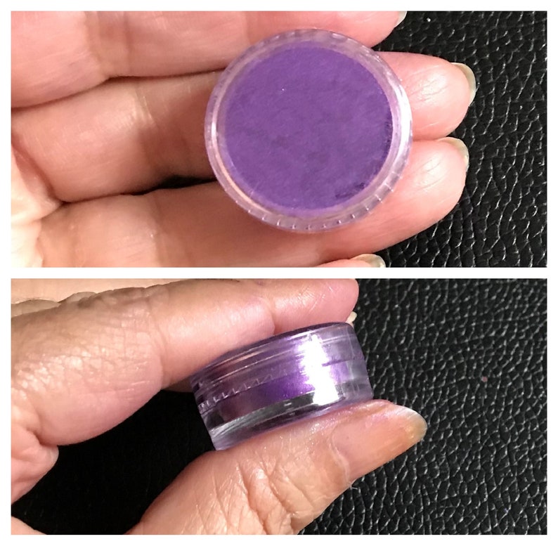 May include: A small clear plastic jar containing a purple iridescent pigment. The jar is being held in a hand.