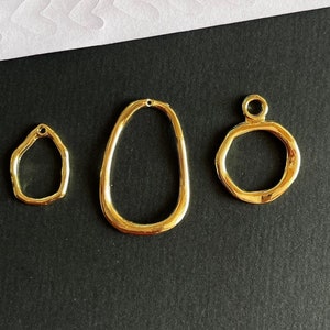 Op de afbeelding: Drie goudkleurige metalen sieradenbevestigingen. De bevestigingen zijn in de vorm van een ovaal, een cirkel en een traan. De bevestigingen zijn allemaal onregelmatig van vorm.