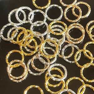 May include: A large pile of silver and gold jump rings. The jump rings are all the same size and have a textured finish.