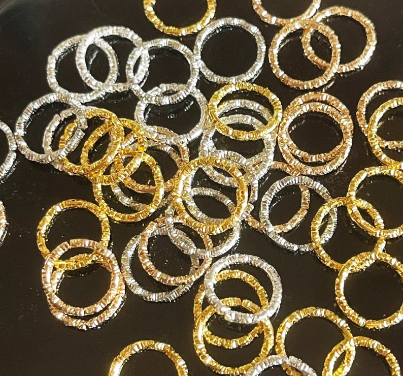 May include: A large pile of silver and gold jump rings. The jump rings are all the same size and have a textured finish.