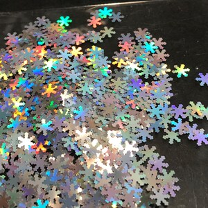May include: A pile of iridescent silver snowflake glitter. The glitter is scattered on a black surface.