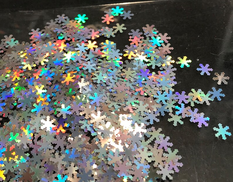 May include: A pile of iridescent silver snowflake glitter. The glitter is scattered on a black surface.