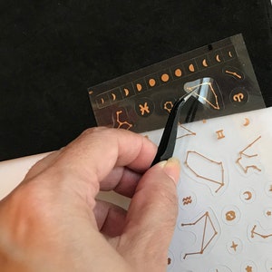 May include: A sheet of gold foil constellation stickers with a pair of tweezers being used to peel off a sticker. The stickers are on a white background and include various constellations, moons, and stars.