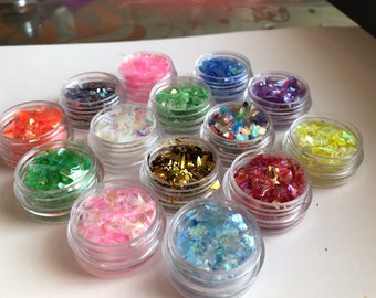 May include: Assortment of 12 small jars filled with iridescent glitter flakes in various colors, including gold, red, green, blue, and pink. The jars are clear plastic with silver lids.