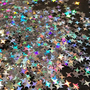 May include: A close-up image of iridescent silver glitter stars scattered on a black surface. The stars are various sizes and shapes, creating a shimmering and festive effect.