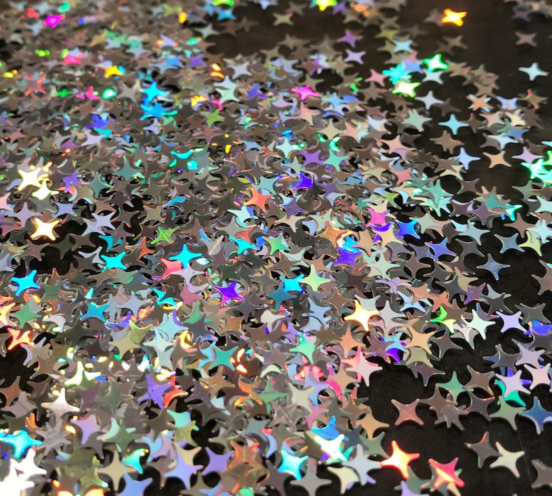 May include: A close-up image of iridescent silver glitter stars scattered on a black surface. The stars are various sizes and shapes, creating a shimmering and festive effect.