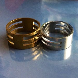 May include: Two rings, one gold-toned and one silver-toned, are displayed. Each ring has a cylindrical band with three horizontal cut-out sections. The gold ring is on the left, and the silver ring is on the right, both set against a blue background.