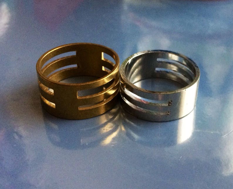 May include: Two rings, one gold-toned and one silver-toned, are displayed. Each ring has a cylindrical band with three horizontal cut-out sections. The gold ring is on the left, and the silver ring is on the right, both set against a blue background.