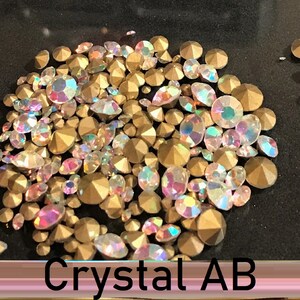 May include: A close-up of a pile of iridescent gold and clear rhinestones. The rhinestones are scattered on a black surface. The text "Crystal AB" is visible at the bottom of the image.