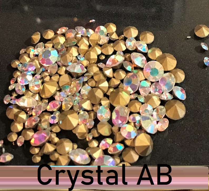 May include: A close-up of a pile of iridescent gold and clear rhinestones. The rhinestones are scattered on a black surface. The text "Crystal AB" is visible at the bottom of the image.