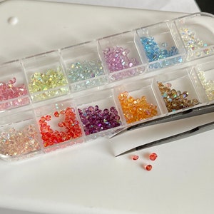 Op de afbeelding: Een doorzichtige plastic opbergdoos met twaalf afzonderlijke vakjes gevuld met kleine, gefacetteerde strass-steentjes in de kleuren roze, geel, blauw, oranje, paars en transparant, vergezeld van een zwarte metalen pincet.