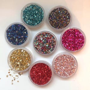 Könnte beinhalten: Sieben kleine, runde, durchsichtige Kunststoffbehälter gefüllt mit zerkleinerten, metallisch glänzenden dekorativen Glassplittern in verschiedenen Farben wie Türkis, Dunkelblau, Gold, Rot, Pink, Roségold und einem bunten, irisierenden Mix, kreisförmig angeordnet.