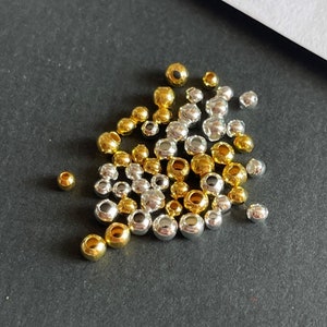 Op de afbeelding: Een verzameling kleine, metalen kralen in goud- en zilverkleuren. De bolvormige kralen variëren in grootte, verspreid over een donkergrijze ondergrond. Ideaal voor het maken van sieraden of knutselprojecten.