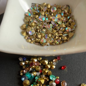 May include: A bowl filled with iridescent gold rhinestones, with a scattering of multi-colored rhinestones on a black surface.