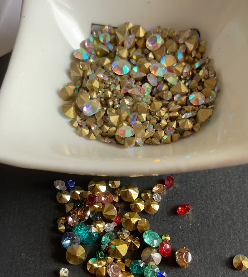 May include: A bowl filled with iridescent gold rhinestones, with a scattering of multi-colored rhinestones on a black surface.