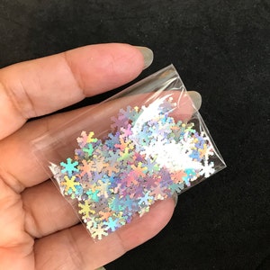 May include: A small clear plastic bag filled with iridescent snowflake-shaped glitter. The glitter is a mix of silver, pink, and blue.