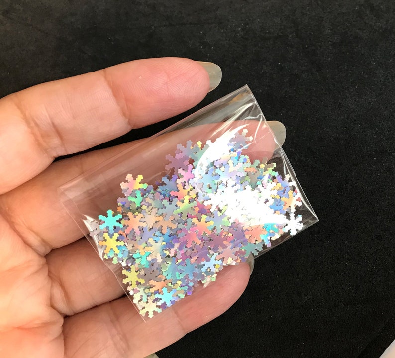 May include: A small clear plastic bag filled with iridescent snowflake-shaped glitter. The glitter is a mix of silver, pink, and blue.