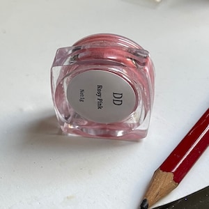 May include: A small, clear jar with a pink cream inside. The jar has a white label with the text "DD Rosy Pink Net:1g".