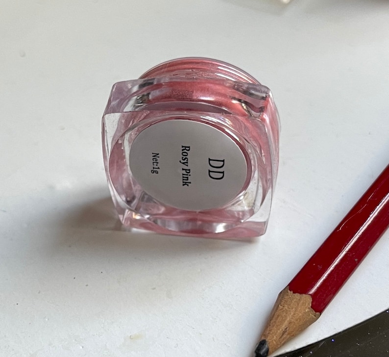 May include: A small, clear jar with a pink cream inside. The jar has a white label with the text "DD Rosy Pink Net:1g".