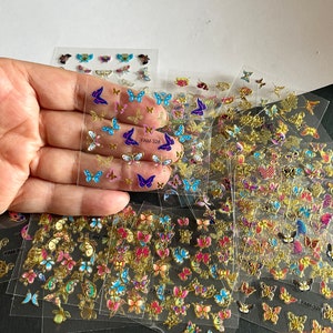 May include: A sheet of clear adhesive nail stickers with colorful butterfly designs. The stickers feature butterflies in various colors, including blue, purple, pink, and gold. The stickers are arranged in rows and columns on a clear sheet. The sheet is labeled "FAM-326".