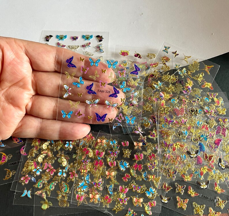 May include: A sheet of clear adhesive nail stickers with colorful butterfly designs. The stickers feature butterflies in various colors, including blue, purple, pink, and gold. The stickers are arranged in rows and columns on a clear sheet. The sheet is labeled "FAM-326".