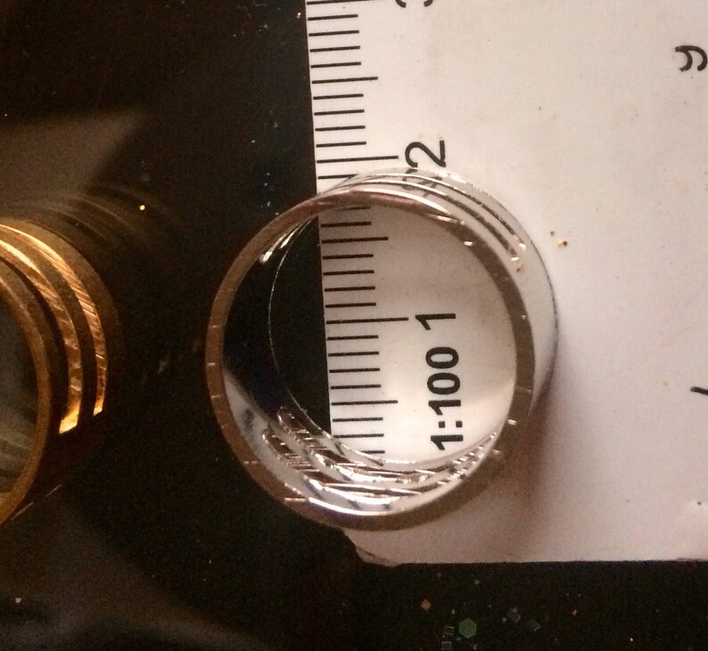 May include: Close-up of a silver ring with a textured design, next to a ruler. The ring appears to be approximately 0.75 inches in diameter. The ring is on a dark surface.