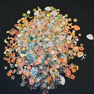 May include: A collection of small, flat decorative sequins in various shapes including stars, moons, flowers, and seashells. The pieces feature a mix of orange, peach, teal, white, and metallic finishes, scattered in a dense, circular arrangement.