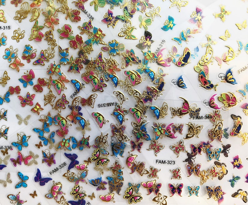 May include: Assortment of colorful butterfly stickers with gold accents. The stickers feature various butterfly designs and colors, including blue, pink, purple, and green. The stickers are arranged in rows on a white background. The stickers are labeled with product codes, including "FAM-328", "FAM-316", "FAM-323", and "M-336".