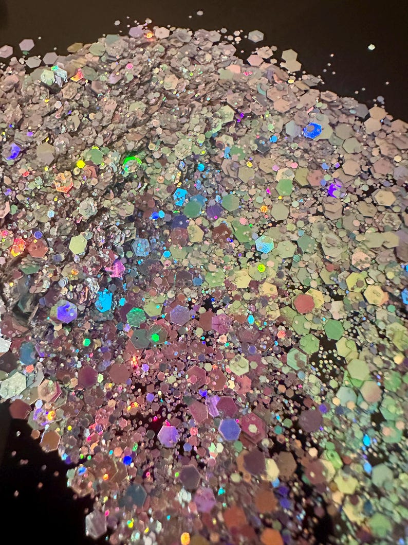 May include: A pile of iridescent glitter with a mix of hexagonal and irregular shapes. The glitter reflects a rainbow of colors, including pink, green, blue, and purple, creating a shimmering effect. Ideal for crafts and decorations.