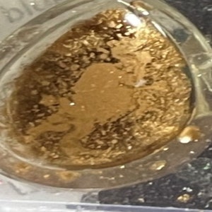 K&ouml;nnte beinhalten: Ein klarer, tropfenf&ouml;rmiger Anh&auml;nger gef&uuml;llt mit Goldflocken. Der Anh&auml;nger hat einen transparenten Rand und ein goldenes Inneres, wodurch ein schimmernder Effekt entsteht. Das Design ist schlicht und elegant, geeignet f&uuml;r Schmuck.