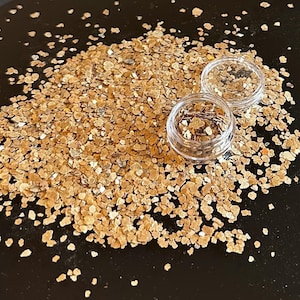 以下が含まれることがあります： 金色のグリッターフレークと2つの小さな透明なプラスチック容器のクローズアップ。グリッターは黒い表面に散らばっています。フレークはメタリックな光沢があり、テクスチャを作成します。