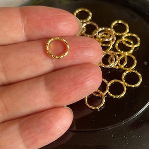 May include: A close-up of a pile of gold-colored metal jump rings. The rings are small and have a textured surface.