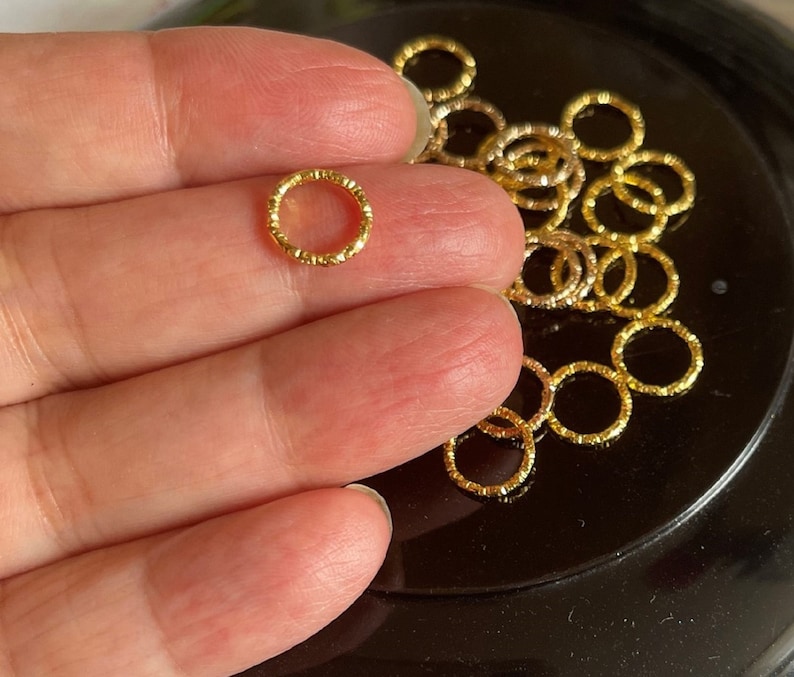 May include: A close-up of a pile of gold-colored metal jump rings. The rings are small and have a textured surface.