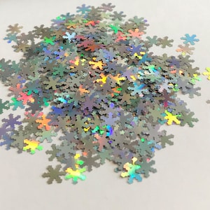 May include: A pile of iridescent silver snowflake-shaped glitter. The glitter is scattered on a white surface.