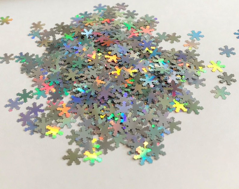 May include: A pile of iridescent silver snowflake-shaped glitter. The glitter is scattered on a white surface.