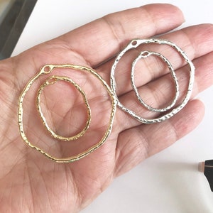 May include: Two metal hoops, one gold and one silver, with a textured finish. The hoops are open and have a small hole at the top for attaching to a chain or other jewelry.