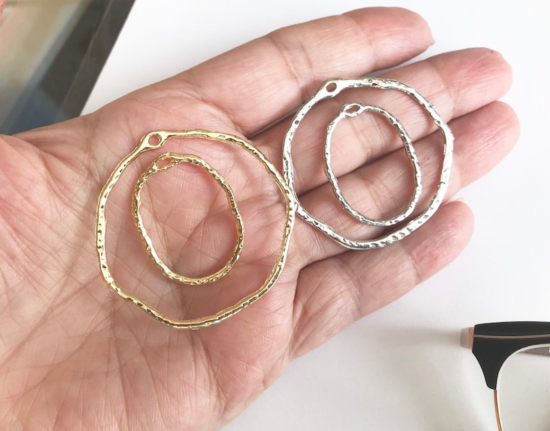 May include: Two metal hoops, one gold and one silver, with a textured finish. The hoops are open and have a small hole at the top for attaching to a chain or other jewelry.