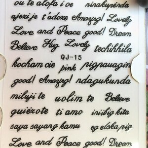 May include: A sheet of clear stickers with black lettering. The stickers feature phrases in different languages, including "Love and Peace", "Amazing!" and "Believe".