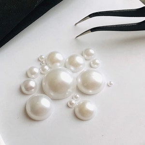 Può includere: Una collezione di decorazioni di perle bianche di varie dimensioni, disposte su una superficie bianca. Le perle sono lisce e lucide, con un leggero bagliore iridescente.