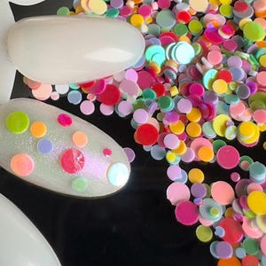 May include: A close-up of colourful round confetti, some with iridescent finishes, scattered on a black surface. Two white nail tips are decorated with colourful polka dots.