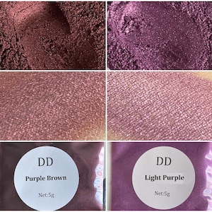 May include: Two small jars of loose eyeshadow pigment. The jar on the left is labeled "DD Purple Brown Net: 5g" and the jar on the right is labeled "DD Light Purple Net: 5g".