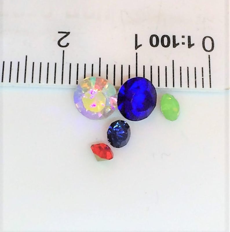 May include: Assortment of five colorful rhinestones, including iridescent, blue, green, and red, on a white surface with a ruler marked in inches.