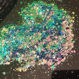 May include: A pile of iridescent glitter in shades of green, blue, pink, and gold. The glitter particles are hexagonal and reflect light, creating a sparkling effect. The glitter is scattered on a dark surface.