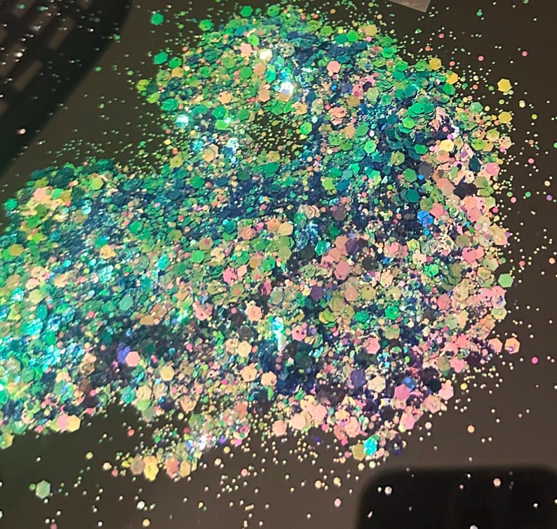 May include: A pile of iridescent glitter in shades of green, blue, pink, and gold. The glitter particles are hexagonal and reflect light, creating a sparkling effect. The glitter is scattered on a dark surface.