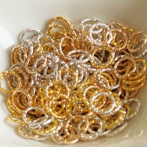 Op de afbeelding: Een stapel gouden en zilveren springringen. De ringen zijn klein en hebben een getextureerd oppervlak.