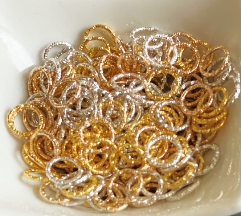 May include: A pile of gold and silver jump rings. The rings are small and have a textured surface.
