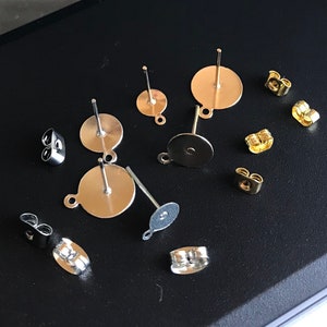 Op de afbeelding: Assortiment van oorbelonderdelen, waaronder zilverkleurige oorbelstiften met ronde achterkanten en goudkleurige oorbelachterkanten.